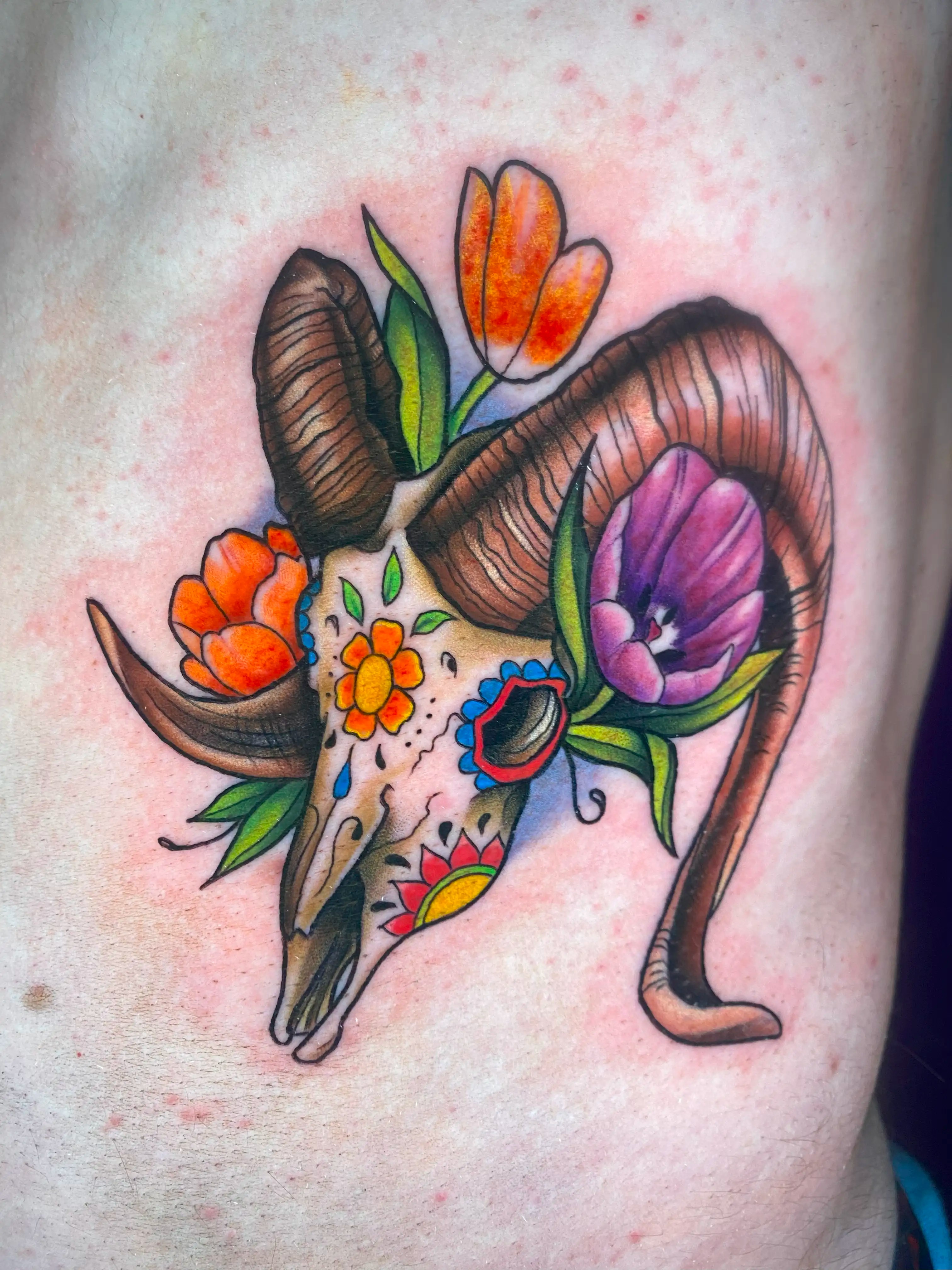 Tattoo of a ram skull with a sugar skull design and flowers around it.
